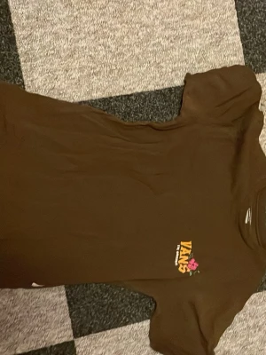 Brun Vans t-shirt med tryck, Small - Säljer en brun t-shirt från Vans i storlek Small. Framsidan har en liten Vans-logga med rosa blomma, medan baksidan har ett stort tryck med dödskalle, palmer och solnedgång. Skön passform och klassisk streetstil, perfekt till jeans eller shorts. Små hål i vänstra sidan.