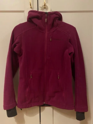 Vinröd fleecejacka/träningsjacka från Norrøna - Säljer en vinröd fleecejacka/träningsjacka från Norrøna i Polartec-material. Jackan har huva, hel dragkedja framtill och två fickor med dragkedja. Perfekt för kyliga dagar och outdoor-äventyr. Broderad logga på bröstet och Polartec-märke på ärmen.