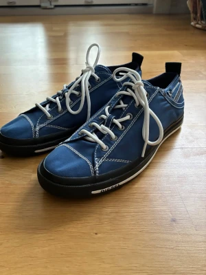 Sneakers från Diesel - Säljer ett par blå sneakers från Diesel med vita kontrastsömmar och vita snören. Svart- och vitrandig sula och en tidlös design. De är tillverkade i Vietnam. Köpte de för 1500 kr. Helt nya och oanvända – perfekta för vardagsbruk.      Kontakta mig gärna om du har frågor eller undrar över något!