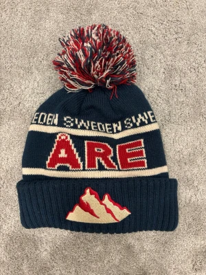 Åre mössa - Stickad mörkblå mössa med stor tofs i rött, vitt och blått. Ribbad kant och klassisk vinterlook, perfekt för kalla dagar.