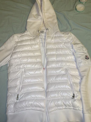 Vit pufferjacka med huva från Moncler - Säljer en clean vit pufferjacka från Moncler med huva och dragkedja framtill. Jackan har quiltad front, mjuka ärmar och två fickor med dragkedja. Klassisk Moncler-logga på ärmen och snörning i huvan för extra stilpoäng.