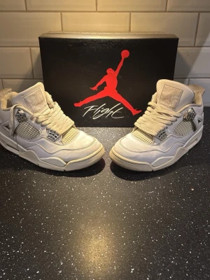 Nike Air Jordan 4 Retro vita sneakers - Säljer ett par klassiska Nike Air Jordan 4 Retro i helvitt skinn med meshpaneler och silverdetaljer. Skorna har snörning, vadderad plös och ikoniska Jumpman-loggor på plös och häl. Perfekta för dig som vill ha en clean och tidlös sneaker med streetkänsla.