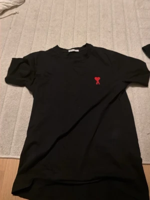 Svart t-shirt med rött hjärta från Ami - Svart t-shirt från Ami med klassisk passform och rund hals. På bröstet sitter ett rött broderat hjärta med bokstaven A. Tillverkad i mjuk bomull som känns skön mot huden. Perfekt för en clean och stilren look.