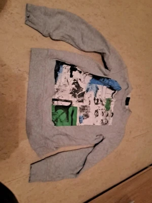 Grå sweatshirt från Warp med tryck - Säljer en grå sweatshirt från Warp i storlek S. Tröjan har ett stort grafiskt tryck framtill i vitt, svart, blått och grönt. Klassisk rund halsringning och långa ärmar. Perfekt för dig som gillar streetwear vibes. I mycket bra skick. Säljs för 120 kr. Tveka inte att kontakta om du är intresserad eller har några frågor! Först till kvarn dom gäller!