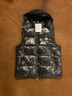Svart dunväst från Moncler - Säljer en svart, glansig dunväst från Moncler med huva och två dragkedjeförsedda fickor. Västen har quiltad design, tydlig Moncler-logga på bröstet och kraftig dragkedja framtill. Perfekt för lager-på-lager under höst och vinter.