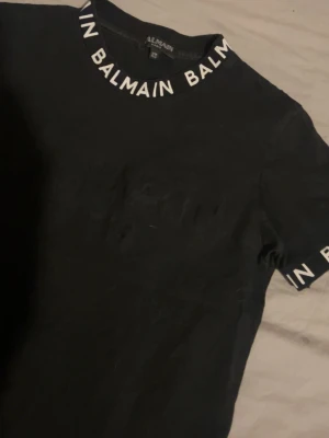 Svart Balmain t-shirt med logga - Säljer en svart t-shirt från Balmain med vit logga runt kragen och ärmsluten. T-shirten har klassisk passform och är tillverkad i mjuk bomull. Enkel men ändå statement med tydlig branding, perfekt till jeans eller shorts.