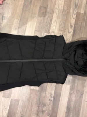 Svart dunväst med huva från Zara - Säljer en svart quiltad dunväst från Zara i storlek M men känns som S . Västen har en stor huva, dragkedja framtill och ribbade muddar i sidorna för extra komfort och passform sanpassad,Perfekt att slänga över en hoodie för en chill och snygg look.