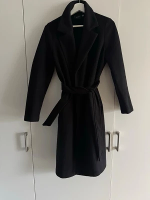 Svart kappa med bälte från vero Moda  - Säljer en svart kappa från Vero Moda med klassisk krage och knytbälte i midjan. Långärmad och rak modell som ger en clean och stilren look. Perfekt för dig som vill ha en enkel men snygg jacka till höst och vinter.  Obs! Kappan är något nopprig. Ca.110cm lång, den kortare varianten från vero Moda 