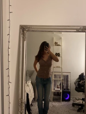 Brun korsettopp med knappar - Säljer en brun korsettopp från Community Co. med snygg v-ringning och rynkade detaljer vid bysten. Toppen har dekorativa knappar hela vägen framtill och är figurnära i modellen. Perfekt att styla med jeans eller kjol för en trendig look.