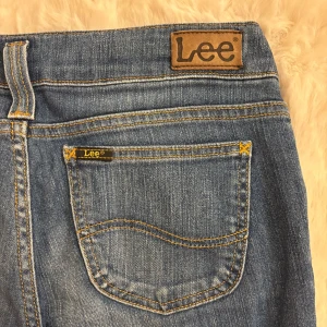 Blå bootcut jeans från Lee - Säljer ett par klassiska blå jeans från Lee med bootcut passform. Tillverkade i tåligt denim med Lee-logga på bakfickan och linningen. Perfekta för en avslappnad och cool stil. Midjemått: 39 cm, innerbenslängd: 76 cm.