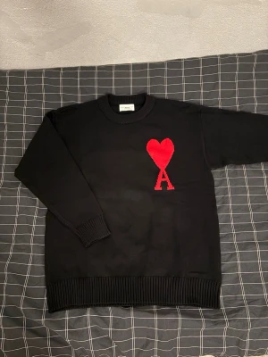 Svart stickad tröja från Ami Paris - Säljer en svart stickad tröja från Ami Paris med deras ikoniska röda hjärta och A på bröstet. Tröjan har rund hals, långa ärmar och ribbade muddar. Perfekt för dig som vill ha en stilren men ändå statement look. Bara testad på,, inga defekter. Hör av dig om du har några frågor.