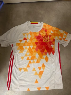 Adidas Spanien matchtröja röd/orange - Säljer en officiell Spanien matchtröja från Adidas med geometriskt mönster i rött, gult och orange. Tröjan har broderad landslagssköld och Adidas-logga på bröstet samt röda ränder på sidorna. Tillverkad i lätt polyester för maximal komfort.