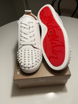 Vita Louboutin sneakers med nitar - Säljer ett par vita sneakers från Christian Louboutin i skinn med ikonisk röd sula och coola nitar på tån. Skorna har snörning och en klassisk låg siluett. Perfekta för dig som vill sticka ut med exklusiv design och edgy detaljer.