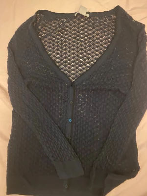 Mörkblå spetskofta från H&M - Säljer en mörkblå kofta från H&M i storlek M. Koftan är stickad med spetsmönster, har v-ringning och knäpps med knappar framtill. Långärmad och lätt transparent, perfekt för lager på lager och en snygg detalj till din outfit.