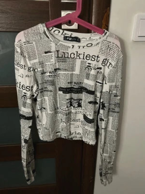 Vit långärmad topp med tidningsmönster  - Säljer en vit långärmad topp från FB Sister med svart tidningsinspirerat print över hela plagget. Modellen är croppad och har en rund halsringning. Materialet är mjukt och stretchigt, perfekt för dig som gillar statement-plagg.