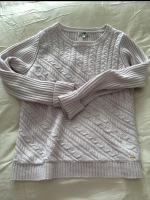 Ljuslila stickad tröja Hampton Republic - Supersnygg ljuslila stickad tröja från Hampton Republic med klassiskt flätmönster och ribbade muddar. Tröjan har rund hals och långa ärmar, perfekt för kyliga dagar. Metallbricka med logga vid nederkanten ger en extra detalj.