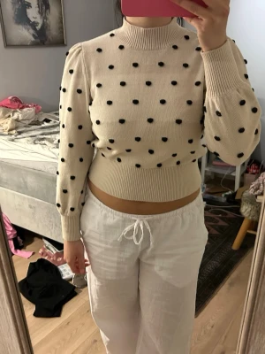 Beige stickad prickig tröja - Superfin beige stickad tröja med svarta prickar och hög krage. Tröjan har lång ärm med lätt puff vid axlarna och bred ribbad mudd i midjan och vid ärmslut. Perfekt för dig som gillar en söt och trendig look.