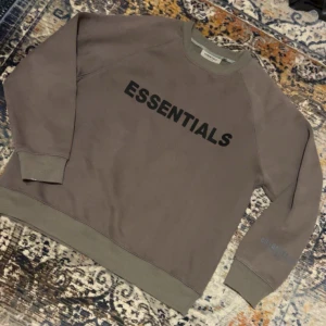 Essentials Fear of God tröja - Säljer en brun sweatshirt från Essentials Fear of God med svart tryck på bröstet och ärmen. Tröjan har ribbade muddar vid hals, ärmslut och nederkant. Perfekt för dig som gillar streetwear och vill ha en clean look. Väldigt fint skick. Storlek Medium 
