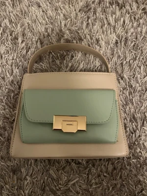 Beige och grön handväska med gulddetalj - Snygg handväska i beige och ljusgrönt med guldfärgat spänne framtill. Väskan har ett topphandtag och avtagbar axelrem, så du kan bära den på flera sätt. Stilren och kompakt modell med ett huvudfack och innerficka. Materialet är slätt och ger en modern look.                               Nypris: 450kr.                                                          Säljs för: 300kr