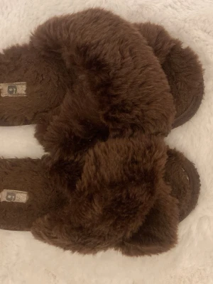 Bruna fluffiga sandaler - Mysiga sandaler i mörkbrunt med fluffig pälsimitation över hela skon. Perfekta för att chilla hemma eller ge din outfit en cozy vibe. Öppen tå och platt sula för maximal komfort.