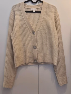 Beige ribbad kofta med strassknappar - Superfin beige ribbad kofta från H&M med glittrande strassknappar. Koftan har v-ringning, lång ärm och är gjord i mjukt stickat material. Perfekt för lager på lager och ger en lyxig touch till din outfit. #abekofta #ribbadkofta #hmstyle #strassknappar #glamdetaljer #v-ringning #stickat #lagerpålager #beigefashion #tidlösstil #vardagslyx #mjuktmaterial #chiclook #scandistyle #modeinspo