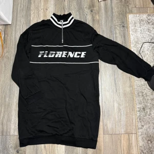 Svart långärmad tröja med half zip - Svart långärmad tröja från H&M Divided med vit text 'FLORENCE' framtill. Tröjan har en randig hög krage med half zip-dragkedja och vita linjer över bröstet.