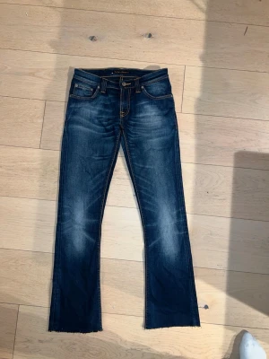 Nudie Jeans Flare Glenn 26/30 - Säljer ett par blå jeans från Nudie med slitna detaljer och råa, avklippta benslut. Jeansen har klassisk femficksdesign, kontrastsömmar och slitna bakfickor med fransar. Materialet är jeans i bomull och passformen är rak med något utsvängda ben.orginal storlek 26/34 men uppklippta