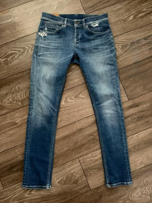 Blå jeans från Dondup - Säljer ett par blå  jeans från Dondup med slitningar vid fickorna och snygg tvätt. Modellen är George. Klassisk femficksmodell med dragkedja och knapp. Jeansen har en smal passform och är tillverkade i mjukt denimtyg för en bekväm känsla.