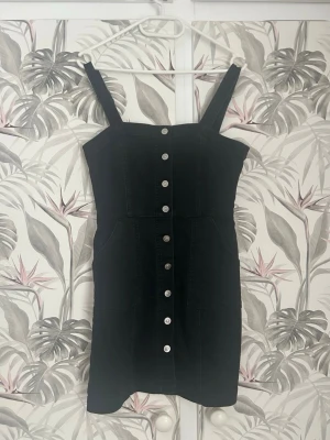 Svart kort jeansklänning med knappar - Säljer en svart kort jeansklänning med breda axelband och silverfärgade knappar framtill. Klänningen har fickor på sidorna och justerbara axelband för perfekt passform. Enkel och stilren modell som är lätt att styla.