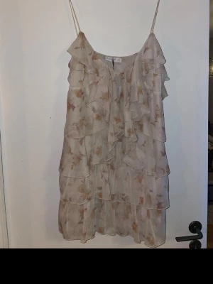 Blommig volangtop från Stradivarius - Säljer en superfin blommig linnetopp från Stradivarius i beige med tunna axelband och flera lager volanger. Toppen har ett skirt, luftigt tyg och ett romantiskt blommönster i ljusa toner. Perfekt för sommarens alla tillfällen!