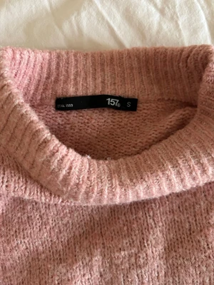 Rosa stickad tröja från 157 - Mysig rosa stickad tröja från 157 med högre ribbad hals och långa ärmar. Tröjan har en relaxed fit och är perfekt för kyliga dagar. Materialet känns mjukt och värmande, och färgen är en fin ljusrosa ton.
