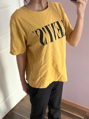 Gul Levi's t-shirt med logga - Säljer en gul t-shirt från Levi's med stor svart logga framtill. Klassisk rund halsringning och korta ärmar. Tillverkad i mjuk bomull som är skön att bära. Har lite fläckar men inget som syns så tydligt 