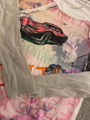 Mesh topp med biltryck och glittriga ärmar - Säljer en unik topp med tryck av en röd sportbil och färgglada grafiska detaljer. Toppen har glittriga, ljusgrå/vita långärmade ärmar och en tight passform. Basen är i ljusa toner med inslag av rosa, orange och blått. Perfekt för dig som gillar statement-plagg! Litet hål vid axeln som ni ser på 5e bilden. Osäker på vilken storlek men skulle gissa på xs/s 