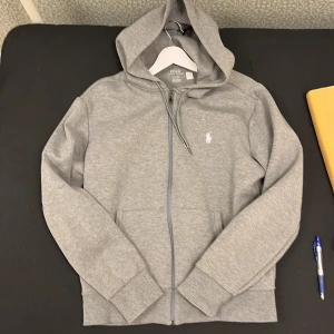 Grå hoodie från Polo Ralph Lauren - Snygg grå hoodie från Polo Ralph Lauren med klassisk logga broderad i vitt på bröstet. Tröjan har huva med snörning, dragkedja framtill och två fickor. Tillverkad i mjuk bomullsmix och har långa ärmar med ribbade muddar. Perfekt för chill dagar.