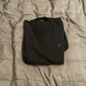 Svart v-ringad tröja från GANT - Svart v-ringad tröja från GANT i storlek L. Tröjan har ett diskret broderat GANT-logo på bröstet och är långärmad med ribbade muddar vid ärmslut och nederkant. Perfekt för dig som gillar stilrena och klassiska plagg.