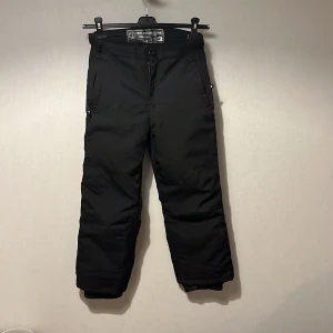 Svarta täckbyxor från Ski Industries - Svarta täckbyxor från Ski Industries med vind- och vattenavvisande material. Byxorna har dragkedjefickor, reflexdetaljer vid bensluten och elastiska muddar. Perfekta för kalla dagar och vinteraktiviteter. Märkta med 'selected edition' och har en clean look.