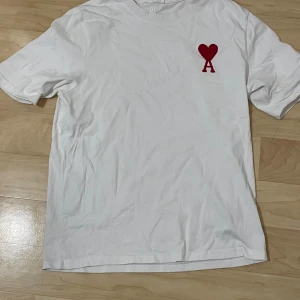 Vit t-shirt från Ami med rött hjärta - Vit t-shirt från Ami Alexandre Mattiussi med klassisk passform och korta ärmar. På bröstet finns ett rött hjärta med bokstaven A. Tillverkad i mjuk bomull, perfekt för en clean och stilren look.