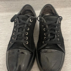 Lanvin med lackad tå - Säljer Svarta sneakers med märket lanvin, snörning och rund tå. Skorna har en snygg kontrast mellan mockaliknande material på ovandelen och en glansig, lackad tå. Metallfärgade öljetter och platt sula ger en clean och stilren look. Perfekta för dig som gillar minimalistisk stil.