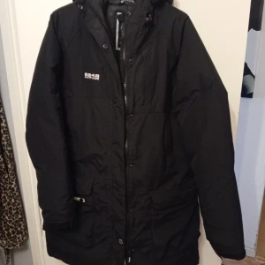 Svart dunjacka från 8848 Altitude - Svart dunjacka från 8848 Altitude med huva och dragkedja framtill. Jackan har quiltad insida, praktiska fickor med dragkedja och en stilren look. Perfekt för kalla dagar och har en längre modell som täcker bra. Snygg logga på bröstet.