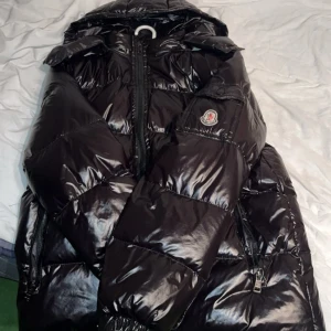Svart pufferjacka från Moncler - Säljer en svart, glansig pufferjacka från Moncler med huva och dragkedja framtill. Jackan har Moncler-logga på ärmen, två sidofickor med dragkedja. Bra skick, M men kan passa som (S). Kom med prisförslag, möts upp i Stockholm kan även frakta 