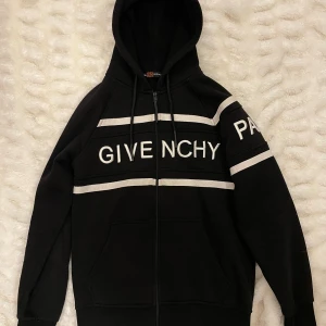 Svart Givenchy hoodie med vita detaljer - Svart hoodie från Givenchy med vita broderade loggor och PARIS-text på ärmen. Tröjan har huva med snörning, dragkedja framtill och två fickor. Snygga vita ränder över bröst och ärmar ger en sportig vibe. Perfekt för dig som gillar streetwear och exklusiva märken.  Det är värda 10 000kr men säljer det för 1000kr 