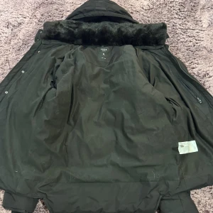 Svart pufferjacka från SMOG XL - Svart pufferjacka från SMOG i storlek XL med snygga detaljer som pälsimitation vid kragen och emblem på ärmen. Jackan har dragkedja, knappar och flera fickor. Materialet är polyester och syntet, perfekt för kalla dagar.