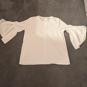 Vit blus med volangärmar - Säljer en vit blus med rund hals och långa ärmar med stora volanger. Blusen har en stilren design och knäppning bak i nacken. 