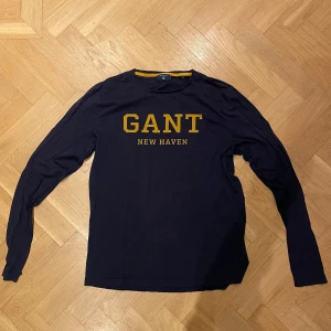 Mörkblå långärmad GANT tröja - Snygg mörkblå långärmad tröja från GANT med gul text 'GANT NEW HAVEN' på bröstet. Klassisk rund hals och mjukt ”T-shirt” tyg. Perfekt för dig som gillar stilrena och sportiga college-vibbar. I bra skick men använd.