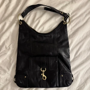 Svart axelväska i skinn  - Cool svart axelväska i äkta skinn från STOCKH LM med mässingsfärgade metalldetaljer och dragkedja. Väskan har en stor ytterficka med spänne framtill och en mindre ficka med dragkedja på baksidan. Snygg och rymlig modell med bred axelrem.