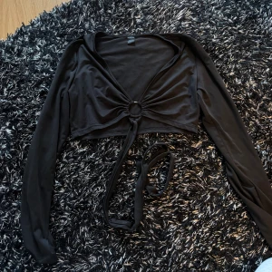 Svart croppad topp med knyte - Säljer en svart croppad bolerotopp med långa ärmar och snygg knytning framtill. Toppen har en ringdetalj i mitten med knyte.  Som ny💗