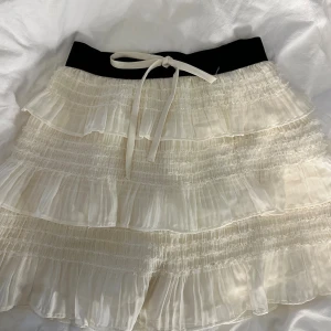 Vit volangkjol med svart midja Pomelo - Supersöt kort volangkjol från Pomelo i vitt med svart midja och knytband framtill. Kjolen har flera lager volanger i ett lätt, skirt material som ger en snygg look! 😻 