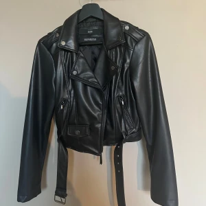 Svart skinnjacka från Cropp - Snygg svart skinnjacka från Cropp med croppad passform, klassisk bikerstil och coola detaljer som bälte i midjan, dragkedjor och knappar. Jackan har långa ärmar och är perfekt för dig som gillar en edgy look.
