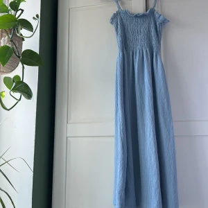 Ljusblå långklänning med smock - Supersöt ljusblå långklänning med smockad överdel, volang vid axlarna och tunna axelband. Klänningen har en flowy passform och är tillverkad i ett krinklat, mjukt tyg som känns luftigt och bekvämt. Perfekt för varma dagar och somriga vibes.