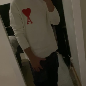 Vit stickad tröja med rött hjärta - Säljer en vit stickad tröja från AMI Paris med ett stort rött hjärta och bokstaven A broderat på bröstet. Tröjan har rund hals och långa ärmar, perfekt för dig som gillar stilrena detaljer med en twist.